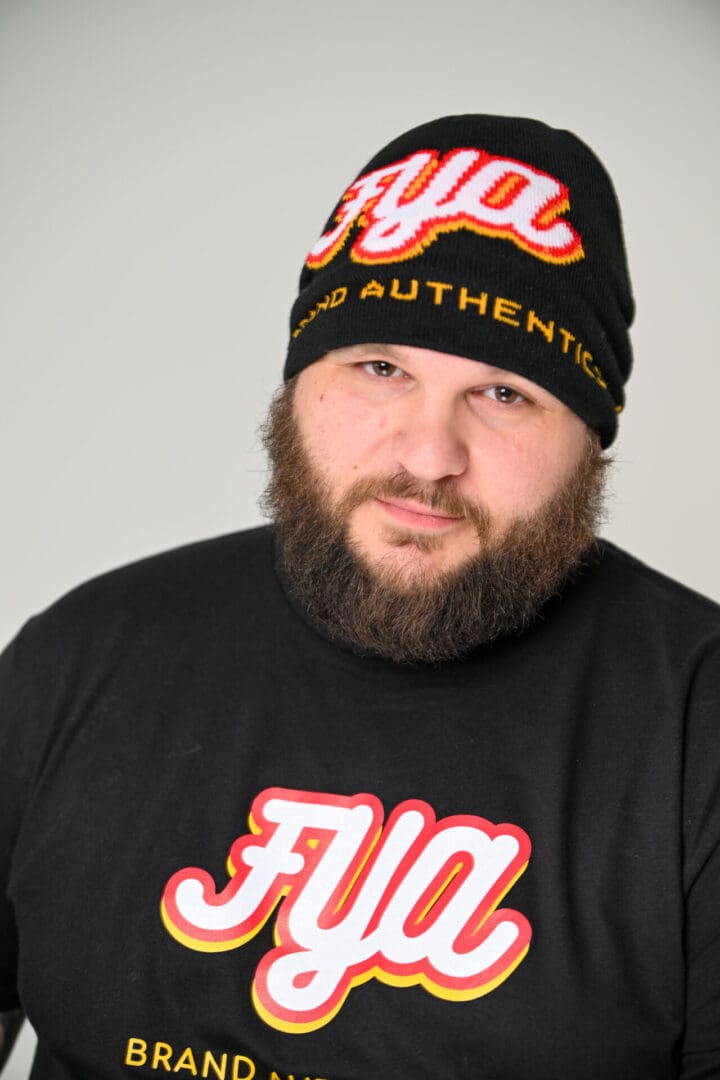 Bearded man wearing a black beanie and black sweatshirt with orange and white text.